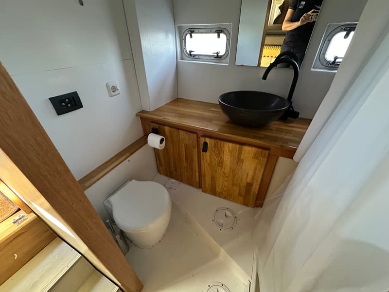 Een witte badkamer met een toilet, houten meubels, een zwarte wasbak en twee patrijspoorten.