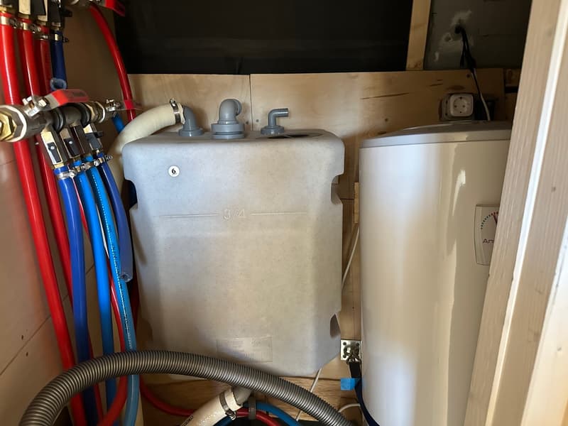 Een detailfoto van een waterboiler en een watertank met diverse leidingen en slangen in een houten compartiment.