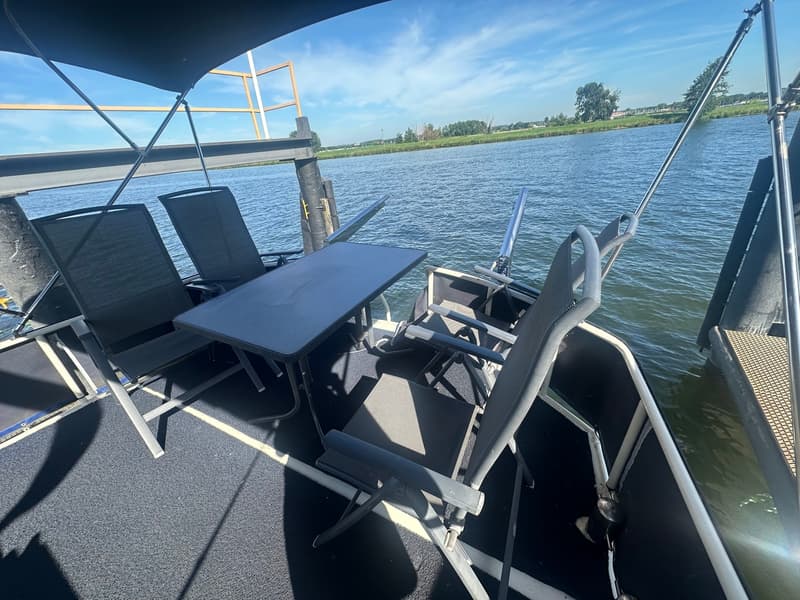 Een dek met een tafel en vier stoelen onder een zonnescherm, met water en een landschap op de achtergrond.