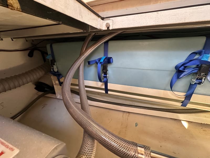 Een lichtblauwe watertank is vastgezet met blauwe spanbanden onder een metalen frame, met slangen op de voorgrond.