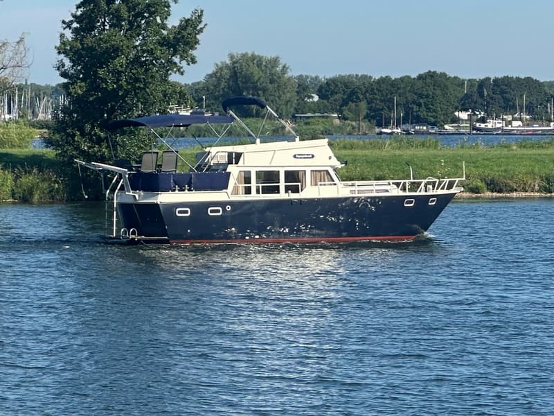 Een blauw-witte motorboot met een rood onderwaterschip vaart op een waterweg met groene oevers en bomen. — Oomen kruiser fly