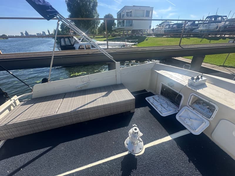 Een achterdek van een boot met een rieten bank, twee open luiken en een lier, met water en gebouwen op de achtergrond.