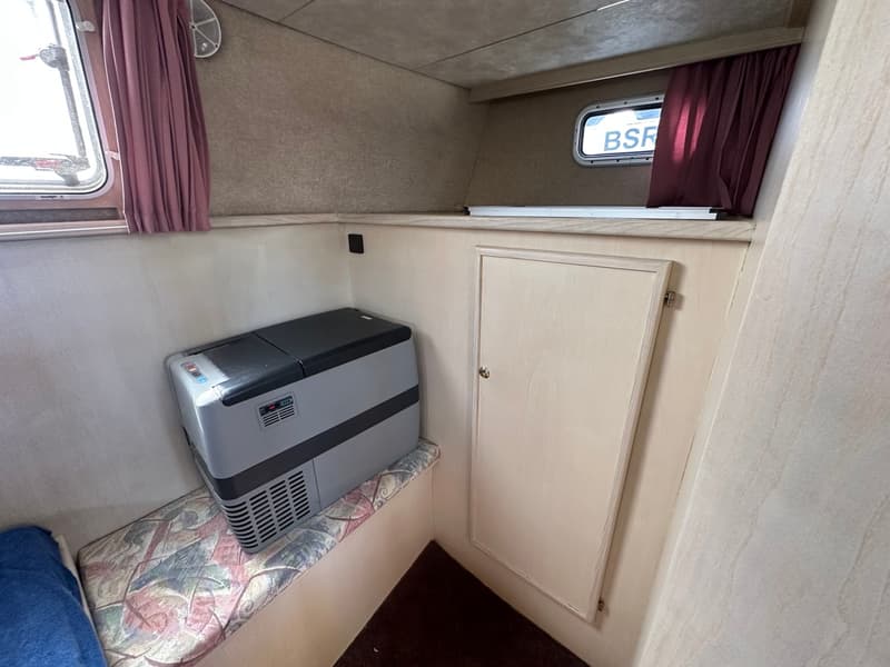 Een interieur van een boot met een grijze koelbox op een gestoffeerde bank, een houten kastje en twee ramen met paarse…