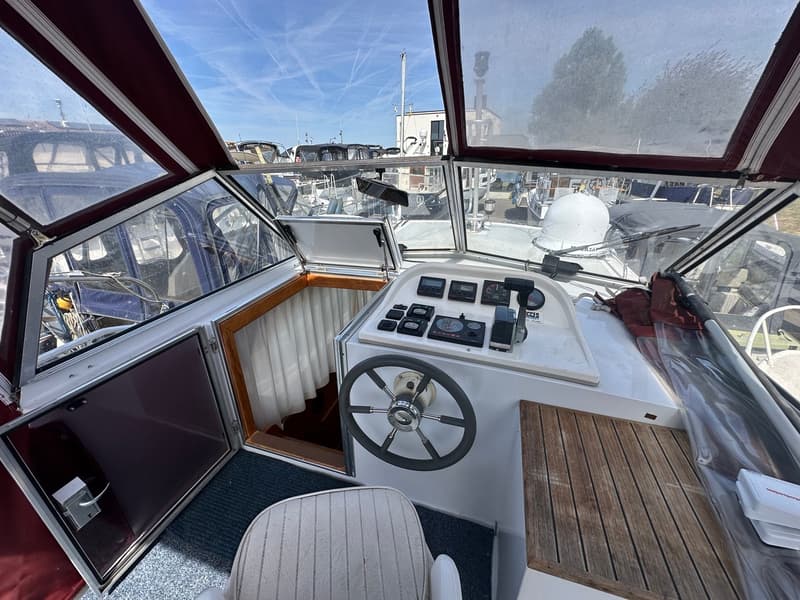 De foto toont de stuurstand van een boot met een stuurwiel, diverse meters en schakelaars, en een houten tafel.