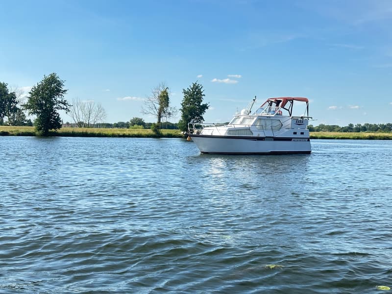 Een witte motorboot met een roodbruine kap vaart op een waterweg met bomen en gras op de achtergrond. — Succes Ultra 500