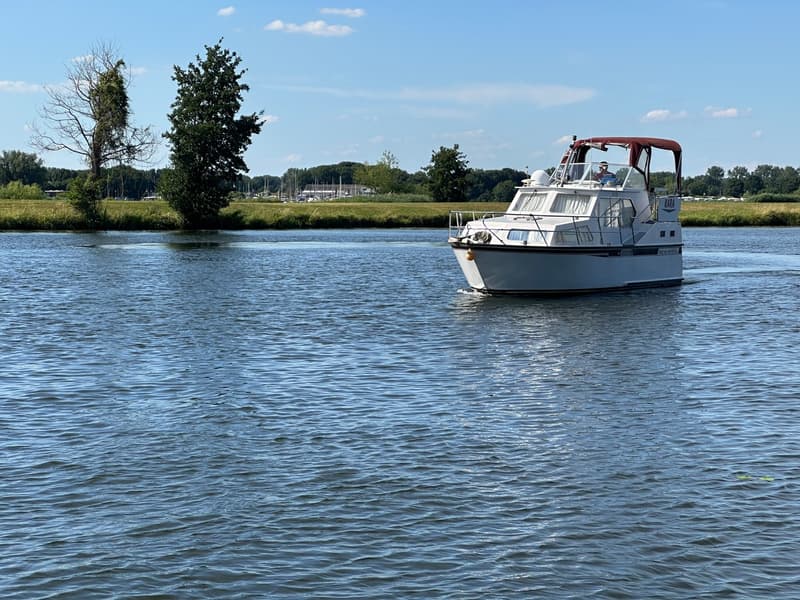 Een witte motorboot met een rood zonnedek vaart op een waterweg, met een groene oever en bomen op de achtergrond.