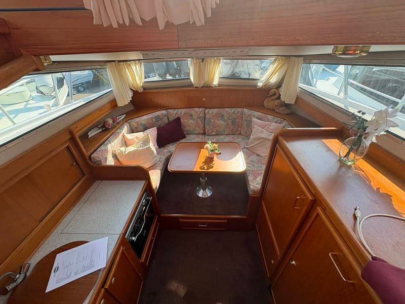 Een interieur van een boot met een U-vormige bank met kussens, een houten tafel en ramen met uitzicht op water en boten.