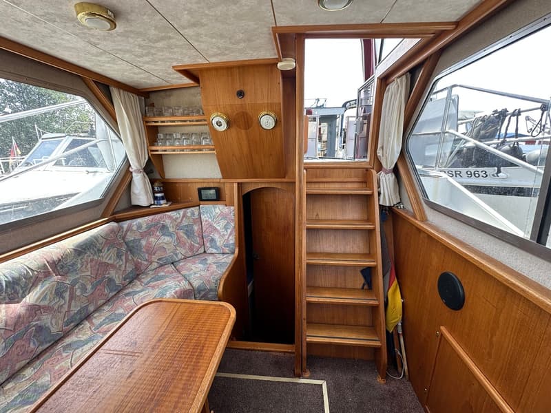 De foto toont het interieur van een boot met een zithoek, een houten tafel, een trap en ramen met uitzicht op een andere…