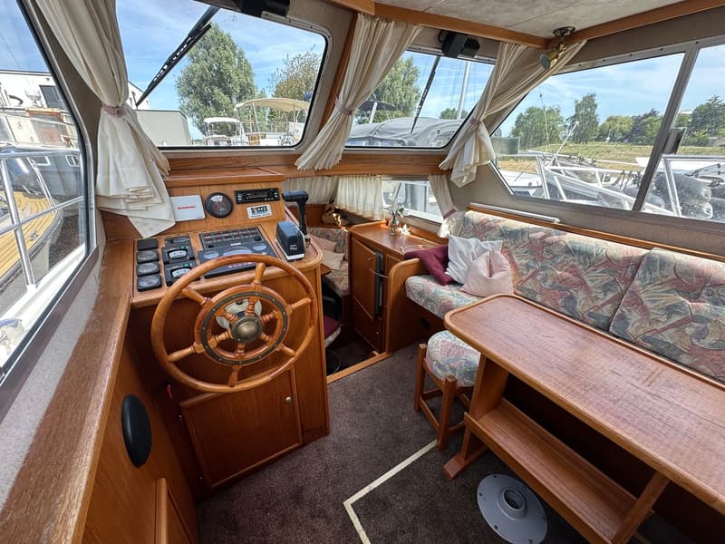 De foto toont het interieur van een boot met een houten stuurwiel, een bank met kussens en een houten tafel.