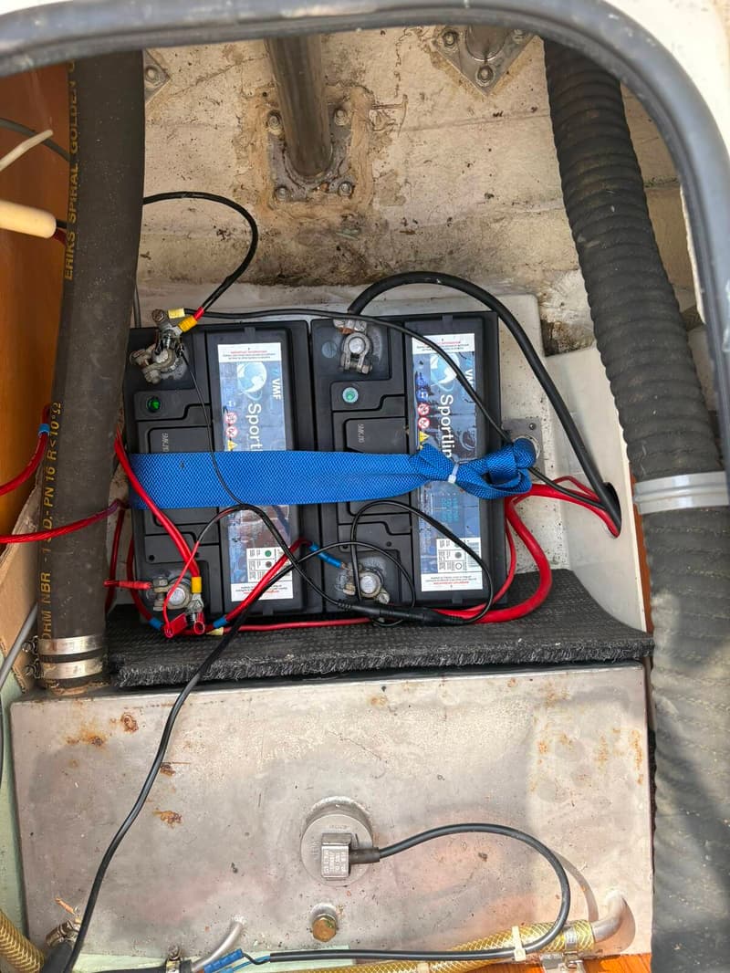 Vier zwarte accu's met blauwe banden en rode en zwarte kabels zijn zichtbaar boven een metalen tank.