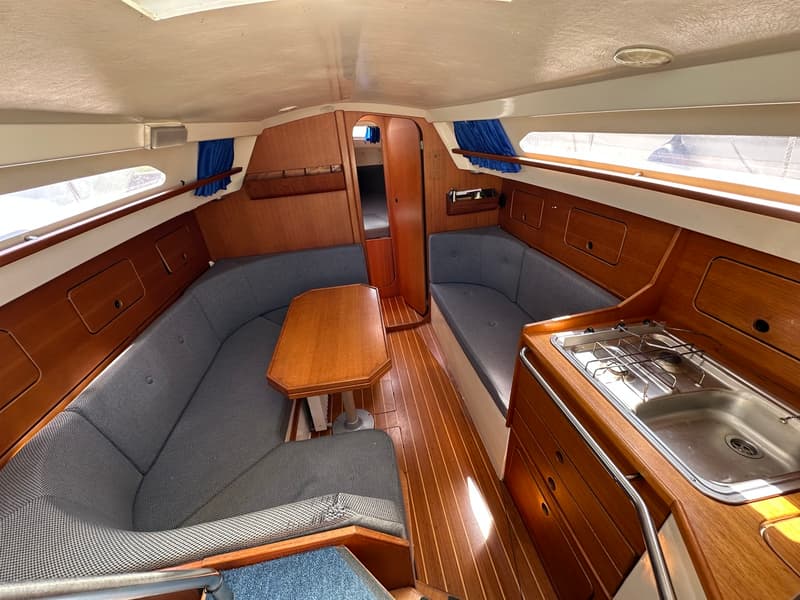 De foto toont het interieur van een boot met houten vloer en wanden, grijze banken, een tafel en een kombuis met fornuis en…