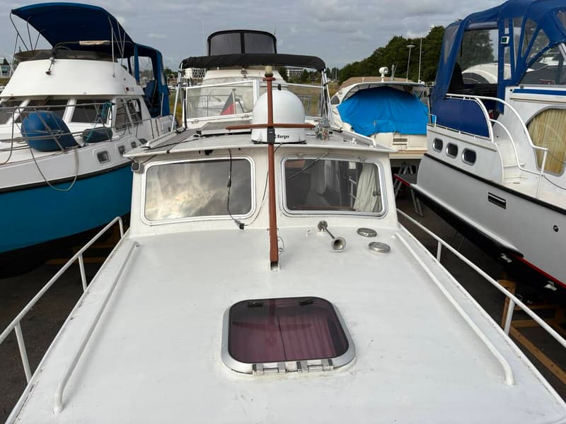 De foto toont het voordek van een witte boot met een luik, twee ramen, een hoorn en een mast, omringd door andere boten.