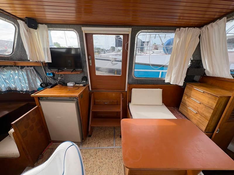 De foto toont het interieur van een boot met een houten plafond, ramen met gordijnen, een televisie, een koelkast, een…