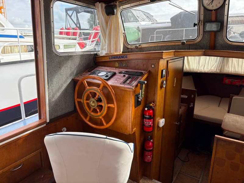 De foto toont het interieur van een boot met een houten stuurwiel, bedieningspaneel, witte stoel en ramen met uitzicht op…