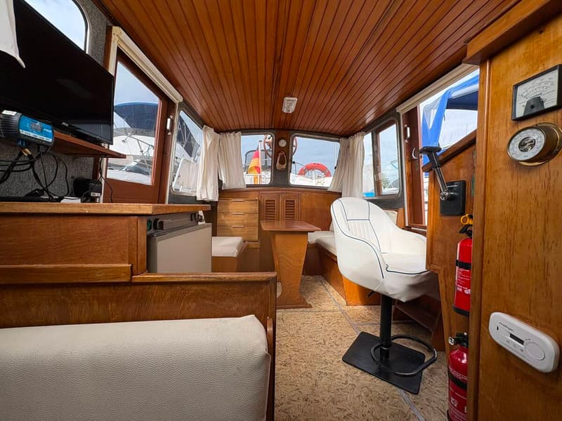 De foto toont het interieur van een boot met houten lambrisering, een witte stoel, banken, een tafel en ramen met gordijnen.