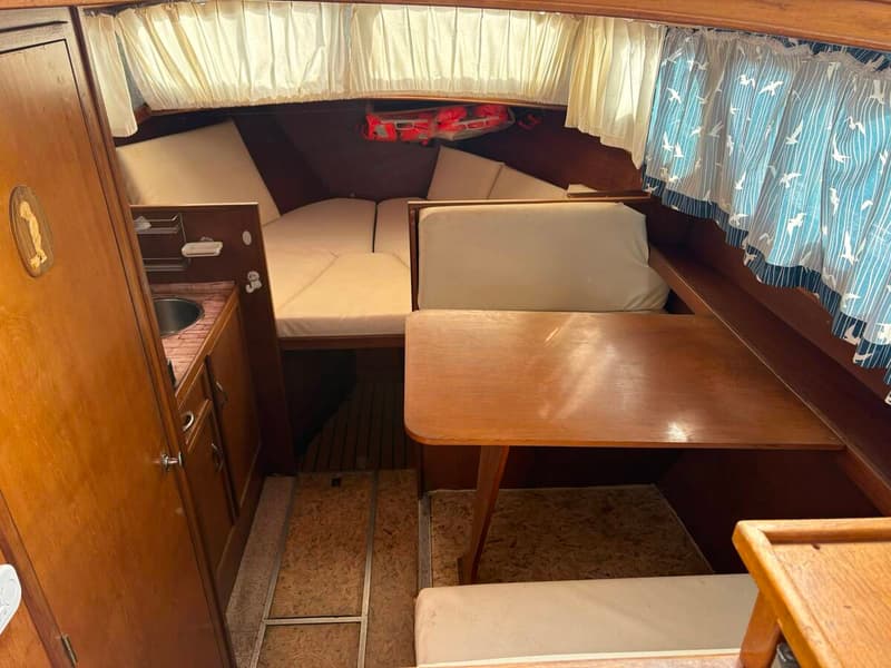 De foto toont het interieur van een boot met een houten tafel, banken met kussens, een bed en een kleine keukenhoek met een…