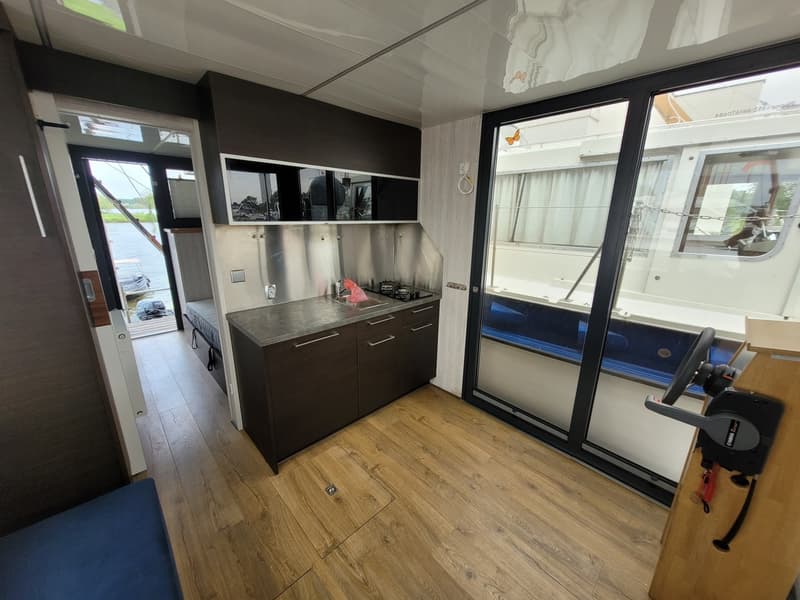 De foto toont een interieur van een boot met een kombuis, houten vloer en een stuurstand naast een glazen schuifdeur.