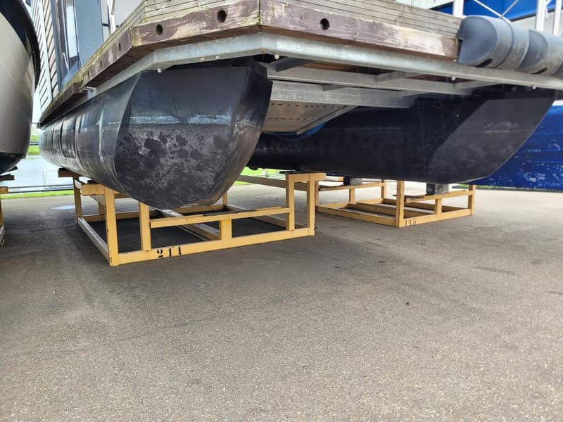 Een pontonboot met twee zwarte drijvers staat op gele bokken op een geasfalteerde ondergrond.