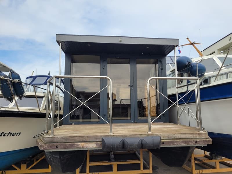 Een woonboot met een houten dek en een glazen opbouw staat op een trailer tussen twee andere boten.