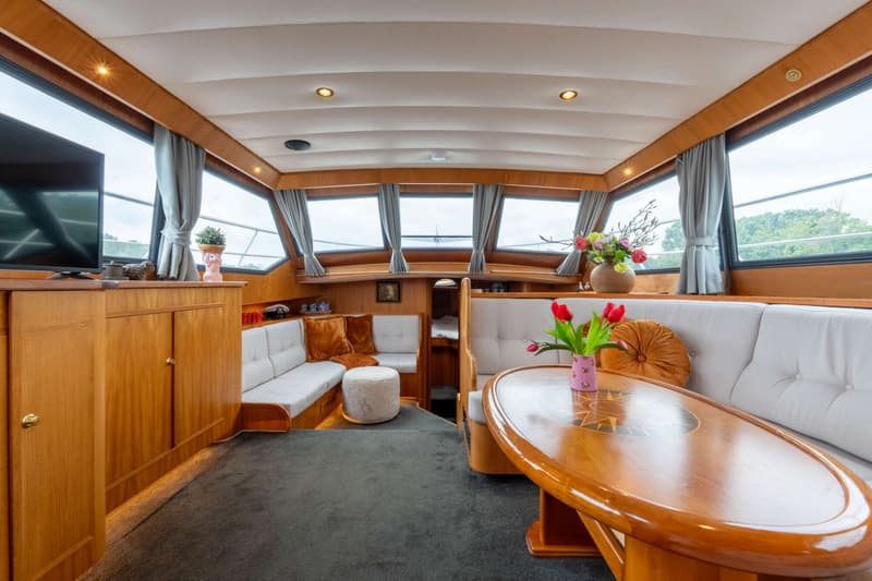 De foto toont het interieur van een boot met houten lambrisering, een grijze vloerbedekking en witte banken.