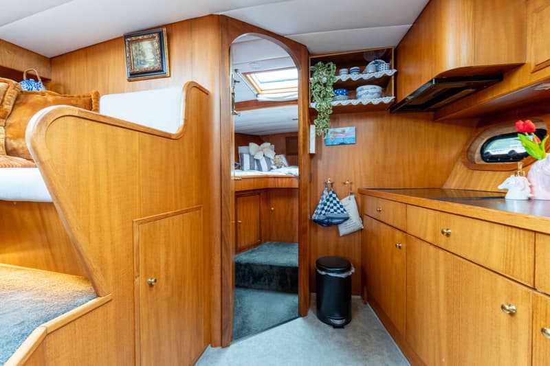 De foto toont een interieur van een boot met houten wanden, een keukenblok, een doorgang naar een slaapruimte en een zithoek.