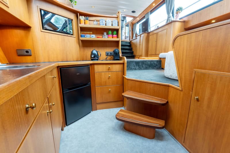 Een houten interieur van een boot met een keukenblok, koelkast, planken, en een trap naar een hoger gelegen dek.