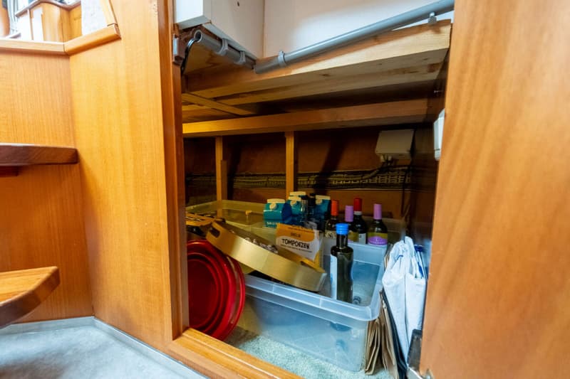 Een opbergruimte met houten wanden en planken, gevuld met flessen, dozen en plastic bakken.
