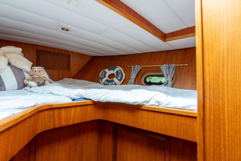 Een kajuit met een bed, houten wanden, een patrijspoort met gordijn, en een reddingsboei-kussen.