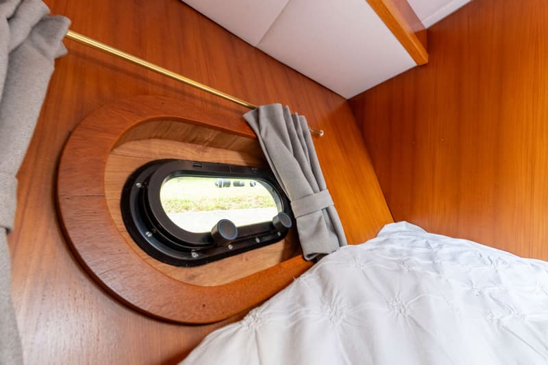 Een patrijspoort met houten omlijsting en een grijs gordijn, naast een wit dekbed.