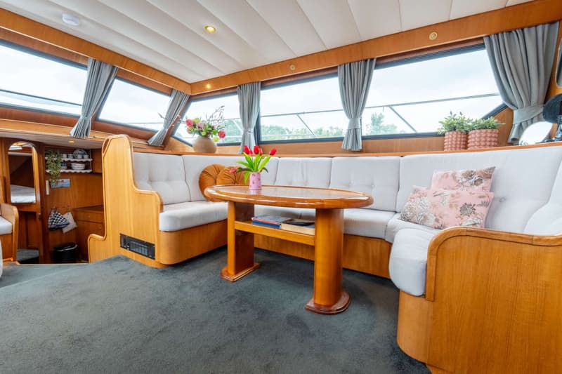 Een interieur van een boot met een houten tafel, grijze banken en ramen met gordijnen.