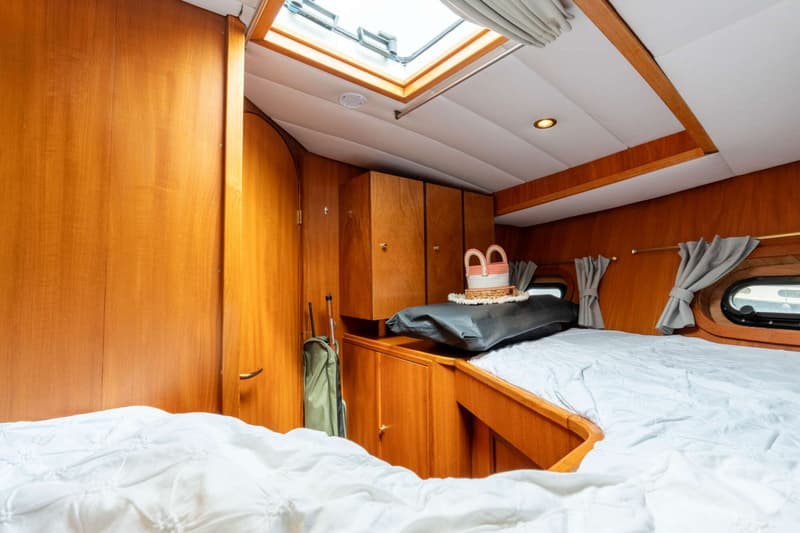 De foto toont een kajuit met een bed, houten kasten, een dakluik en patrijspoorten met gordijnen.