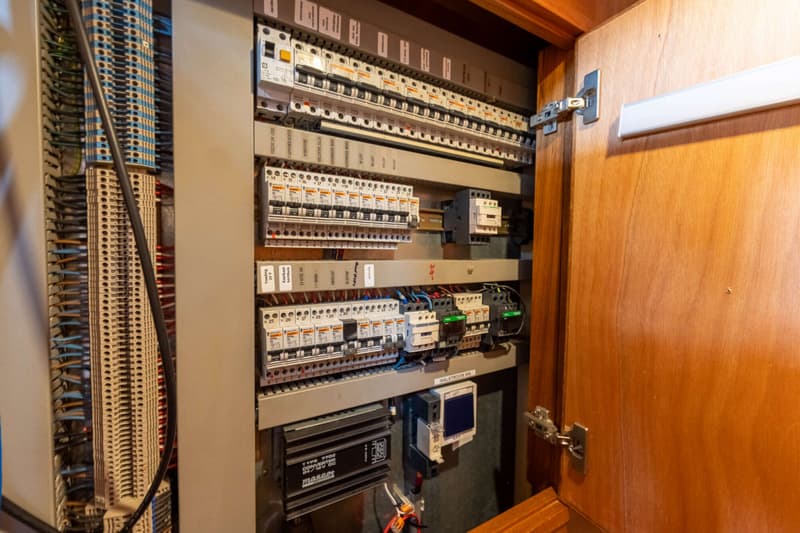 Een open houten kastdeur toont een elektrisch paneel met diverse schakelaars, zekeringen en bedrading.