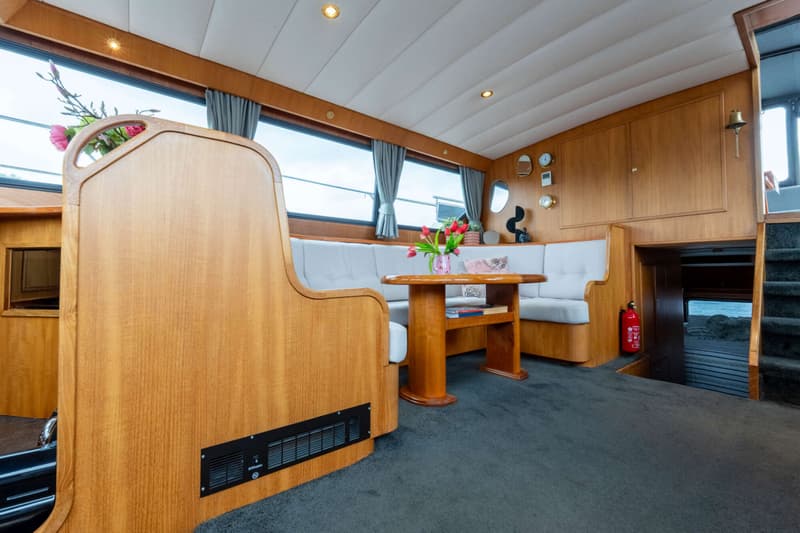 De foto toont het interieur van een boot met een houten tafel, een U-vormige bank, ramen en een trap naar beneden.