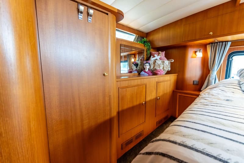 Een kajuit met een bed met gestreepte dekens, houten kasten, een spiegel en een raam met gordijnen.