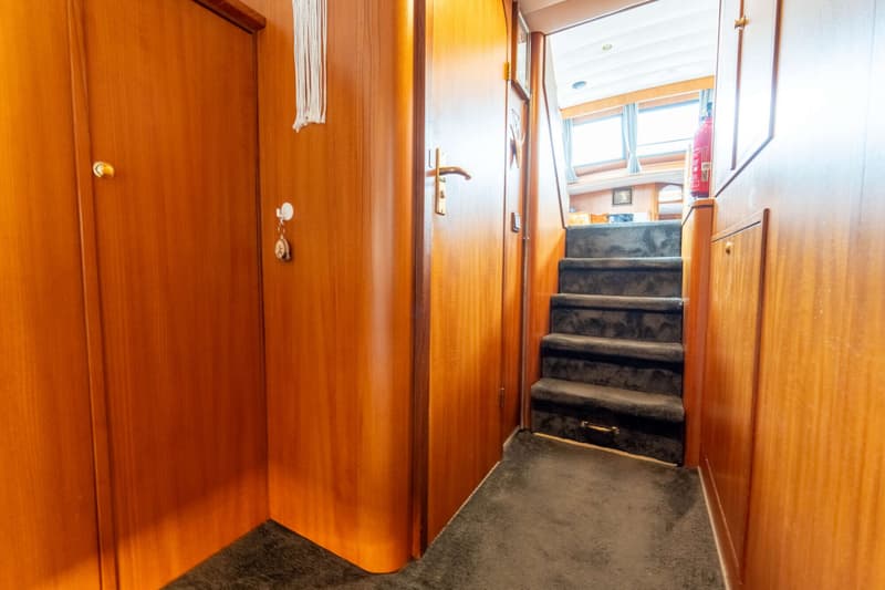 Een houten gang met een donkergrijze trap die naar een hoger dek leidt, met een brandblusser zichtbaar bovenaan de trap.