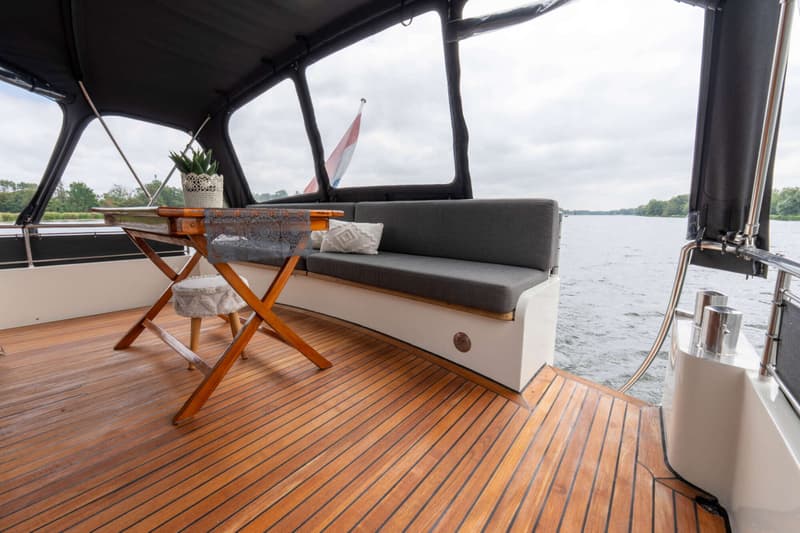 Een houten dek met een tafel, een bank met grijze kussens, en een uitzicht op water en bomen onder een bewolkte lucht.