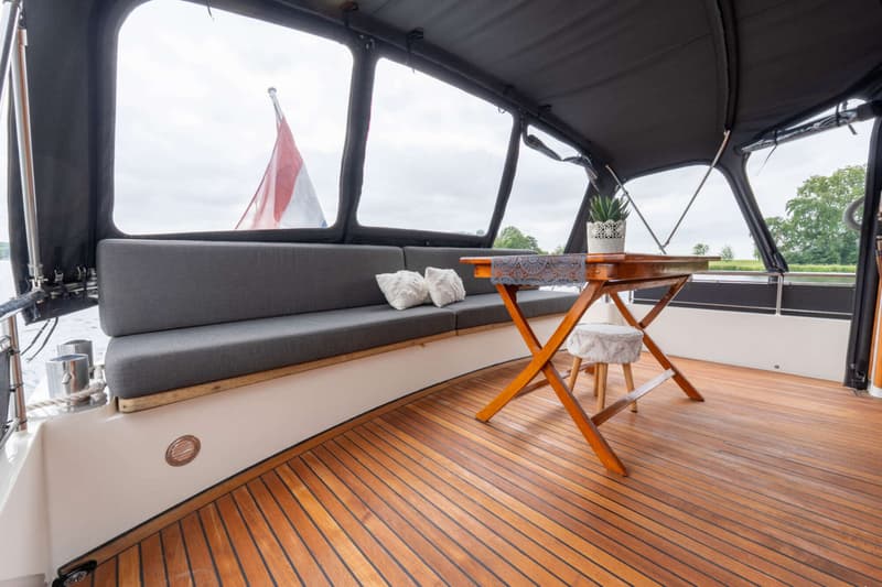 Een houten dek met een bank met grijze kussens, een houten tafel en een Nederlandse vlag in de verte.