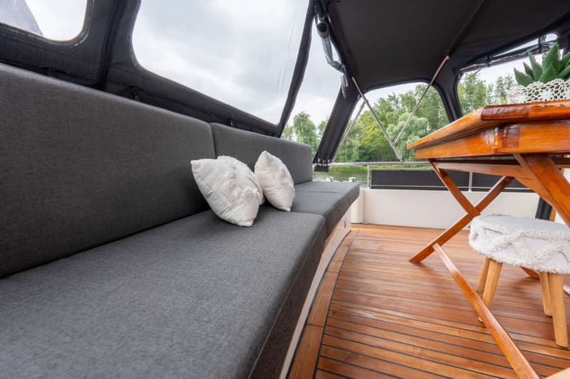 De foto toont een bootinterieur met een donkergrijze bank met twee witte kussens, een houten tafel en een houten vloer.