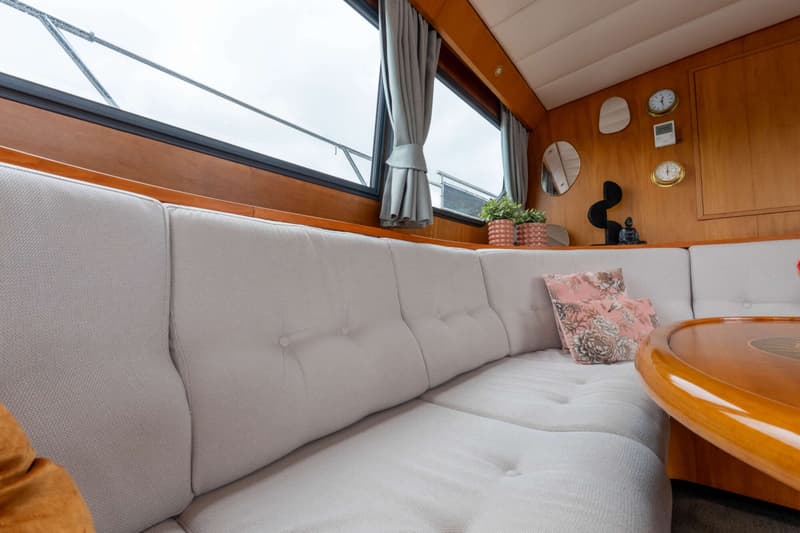 Een interieur van een boot met een lichte bank, houten wanden, ramen met gordijnen en een houten tafel.