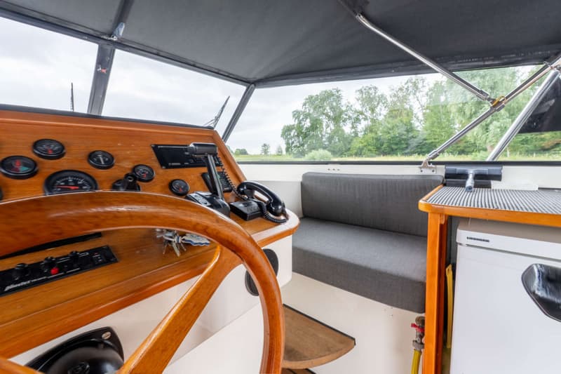 Een houten stuurwiel en dashboard met meters, een bankje, een koelkast en een telefoon in de stuurhut van een boot.