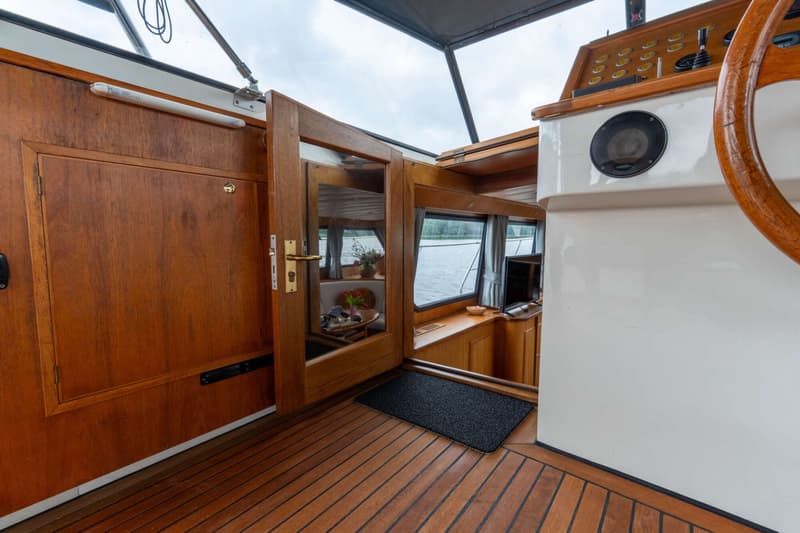 Een interieur van een boot met houten vloer en wanden, een stuurwiel en een open deur naar een kajuit.