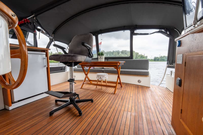 Een houten stuurwiel, een grijze stoel, een houten tafel en een bankje op een houten dek van een boot.
