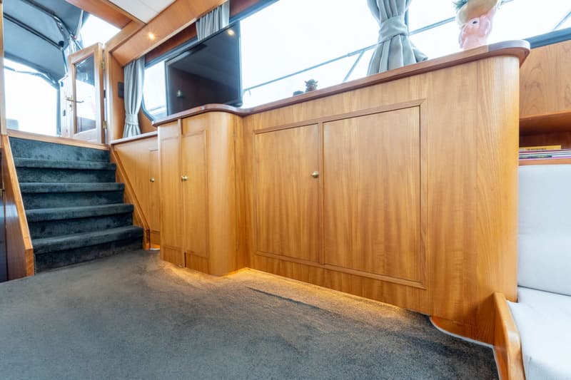Een interieur van een boot met een trap met donkergrijze vloerbedekking, houten kasten en een televisie.