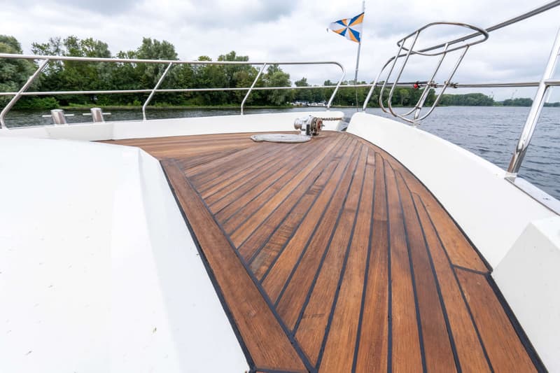 De foto toont het voordek van een boot met een houten dek, witte zijkanten, een reling en een vlag.