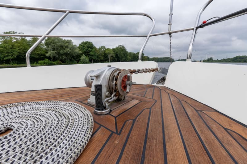 Een ankerlier met ketting, een opgerolde lijn en een houten dek zijn zichtbaar op de boeg van een boot.
