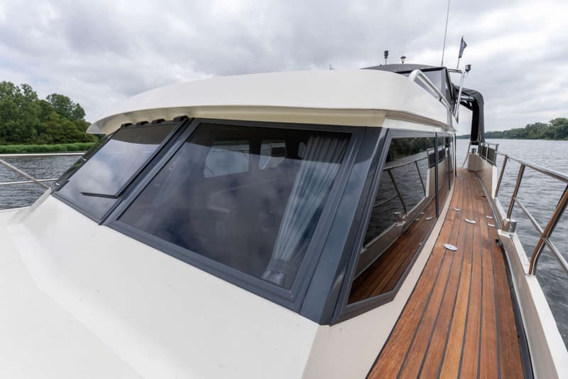 De foto toont de voorkant en zijkant van een boot met een wit dek, houten loopdek en grote ramen met gordijnen.