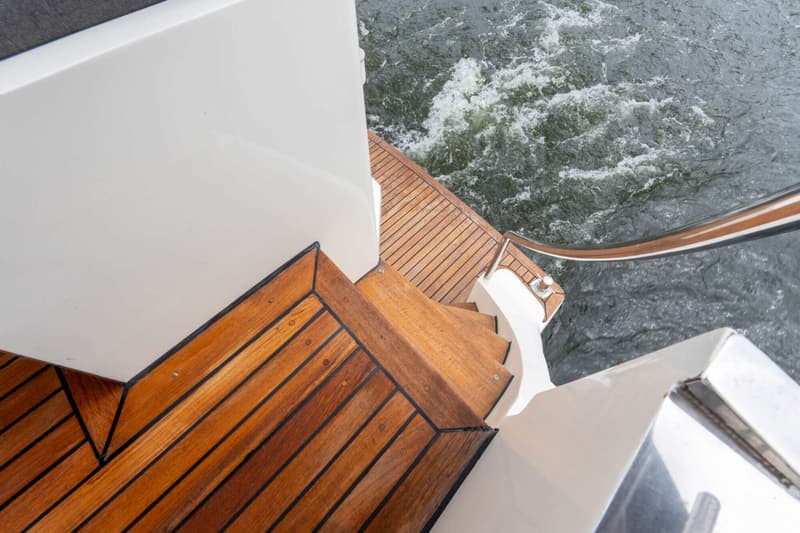Een bovenaanzicht van een houten trap en dek aan de achterzijde van een boot, met water en schuim eromheen.