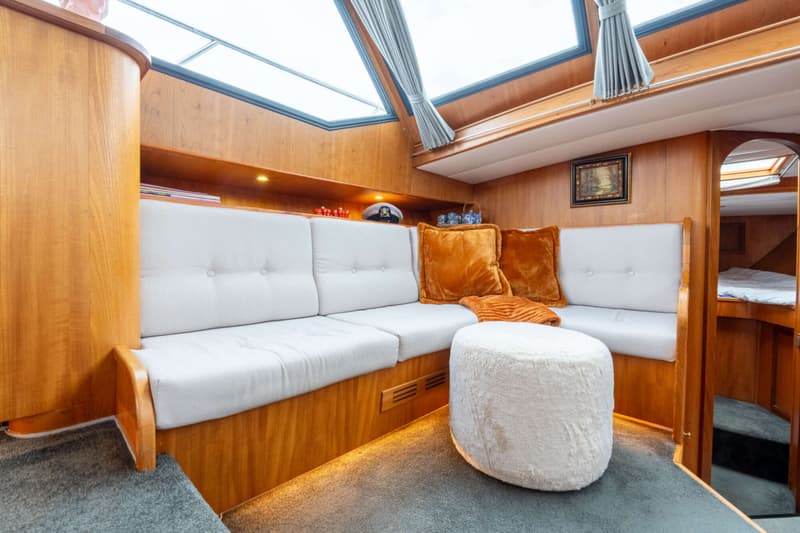 Een interieur van een boot met een L-vormige bank met witte kussens, oranje sierkussens, een witte poef en houten wanden.