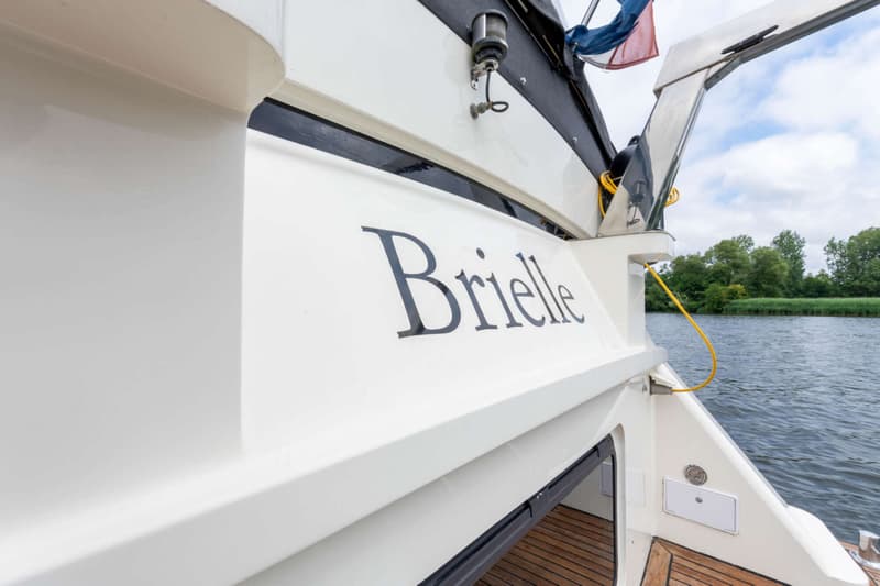 De naam "Brielle" staat in donkere letters op de witte romp van een boot, met een rivier en bomen op de achtergrond.