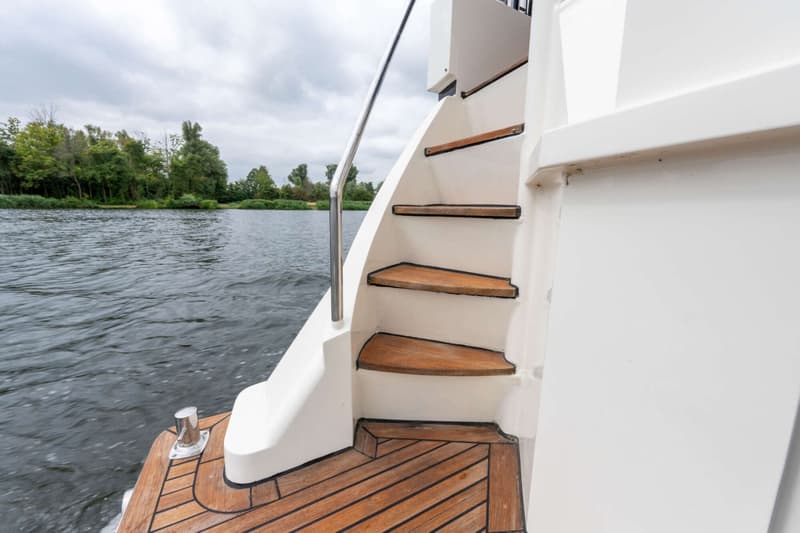 Een witte boot met houten dek en trap, met een stalen reling, vaart op een rivier langs een beboste oever onder een…